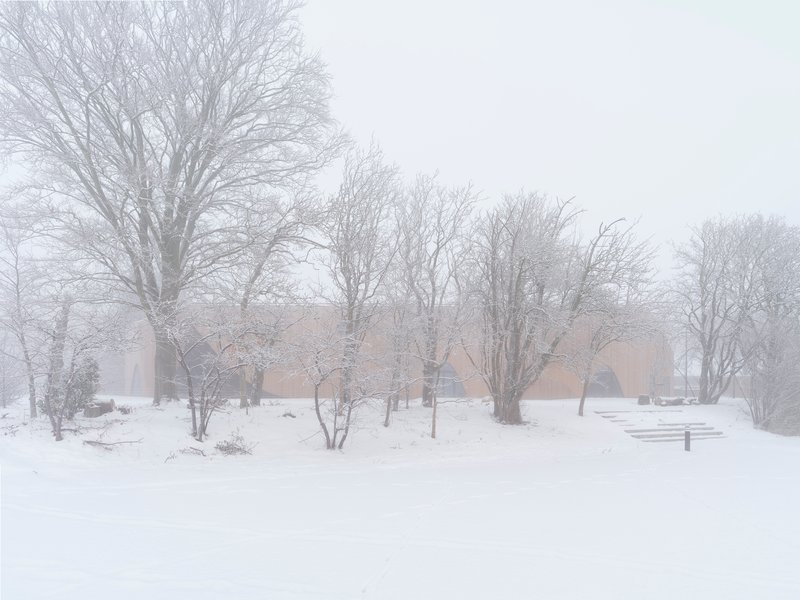 Distant view of the low-slung beige brick structure amid bare winter trees in heavy snow and fog