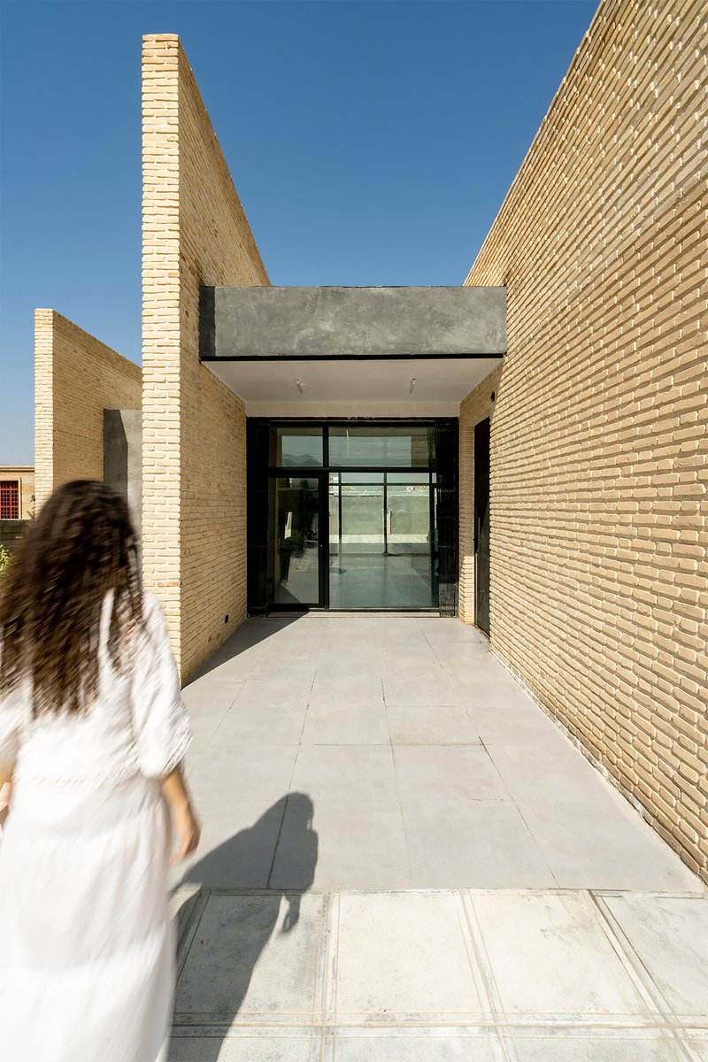 Entrance passageway lined with textured buff brick walls leading to glass doors under a concrete canopy