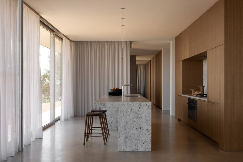 Open kitchen with terrazzo island and timber cabinetry beneath sheer curtains filtering daylight from the courtyard
