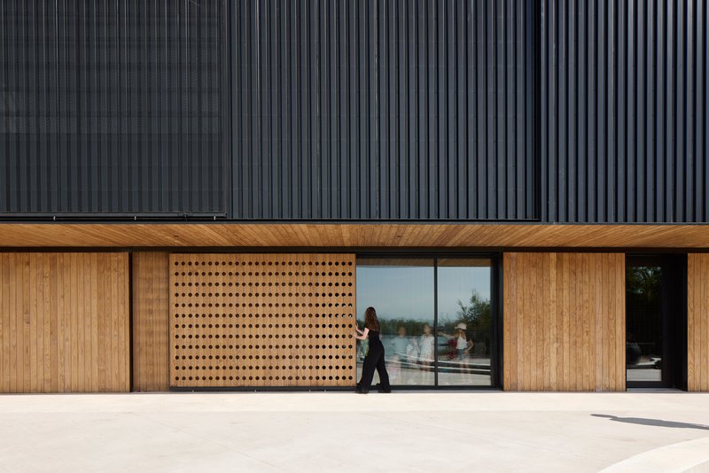 Ground-level facade with perforated timber panels and glazed openings beneath dark corrugated metal cladding as a person walks past