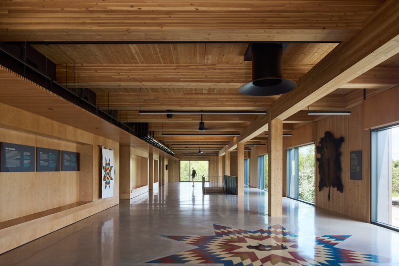Interior gallery space with exposed timber beams and plywood walls displaying a patterned floor installation under linear lighting