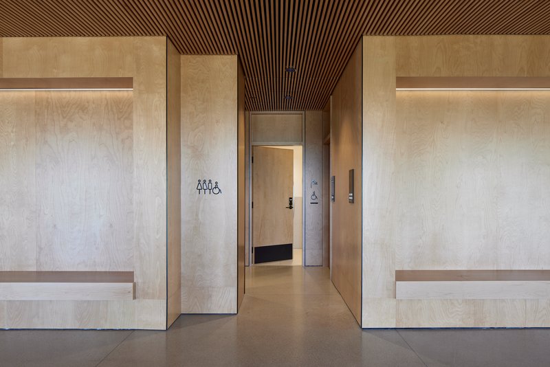 Corridor with light plywood wall panels and integrated benches beneath a slatted wood ceiling