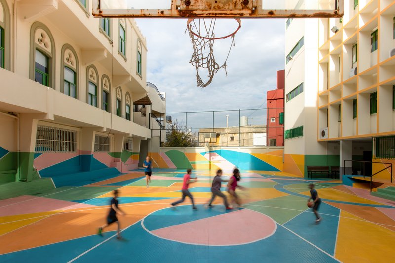 Basketball hoop suspended above the courtyard as children in motion play across the painted surface