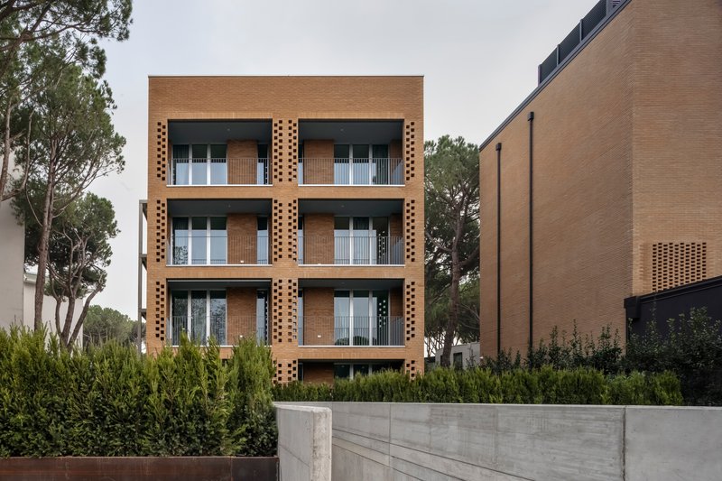 Four-story perforated brick facade with recessed balconies behind a concrete retaining wall and planted hedge