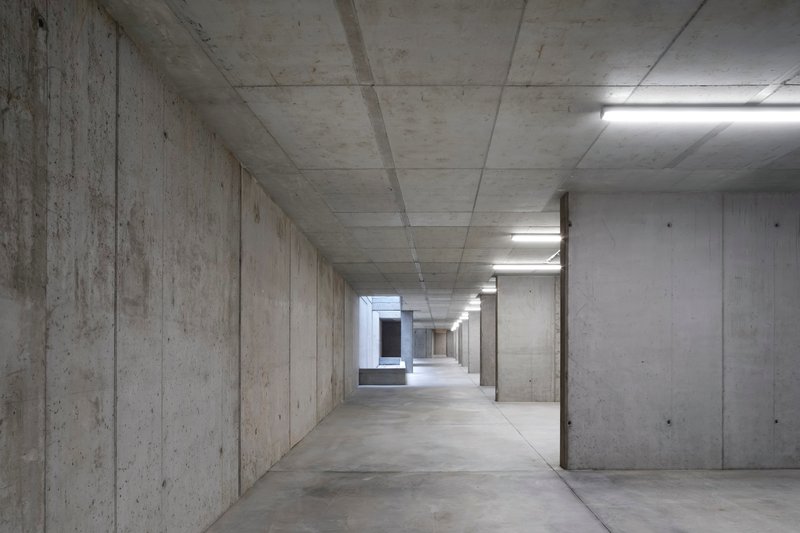 Board-formed concrete corridor with exposed ceiling beams and fluorescent light strips leading to a distant opening