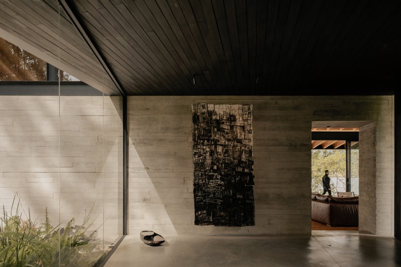 Gallery hallway with board-formed concrete walls, dark timber ceiling, and figure silhouetted in distant doorway