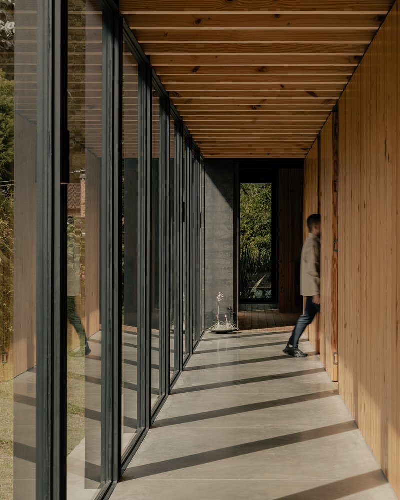 Glazed corridor with person walking on concrete floor beneath exposed timber joists casting shadows