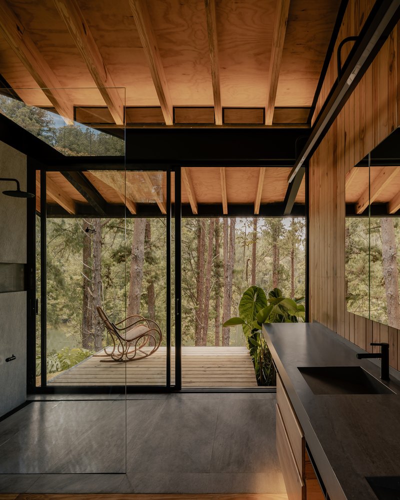 Interior view through floor-to-ceiling glass walls opening to a timber deck with rocking chair among trees