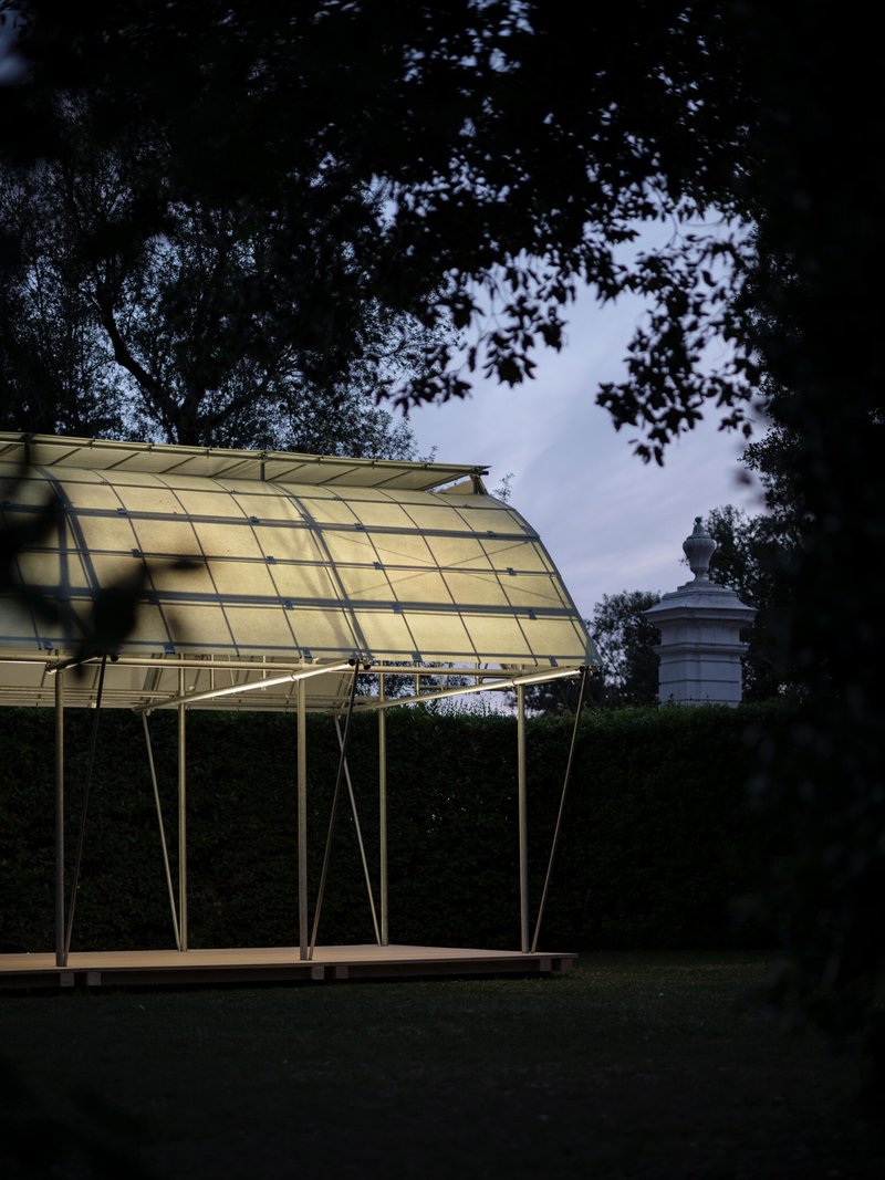 Curved translucent roof structure supported by angled steel legs beneath overhanging trees at dusk