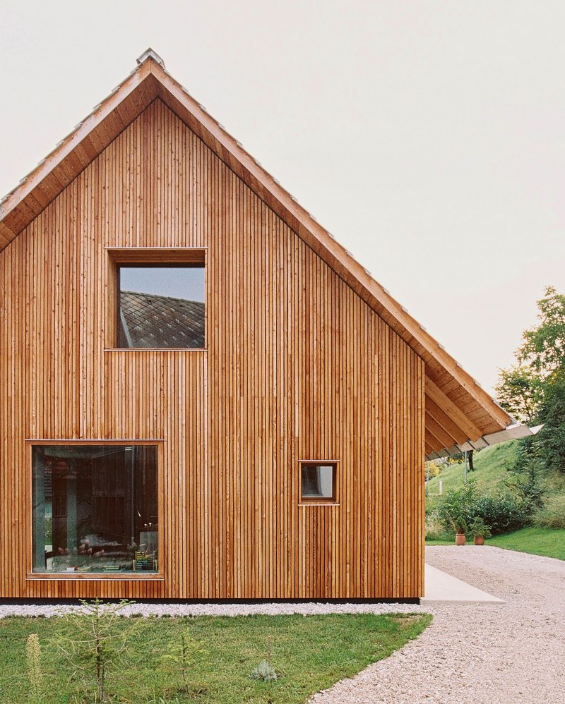 Vertical timber-clad gable facade with square windows flanked by a gravel drive and lawn at dusk