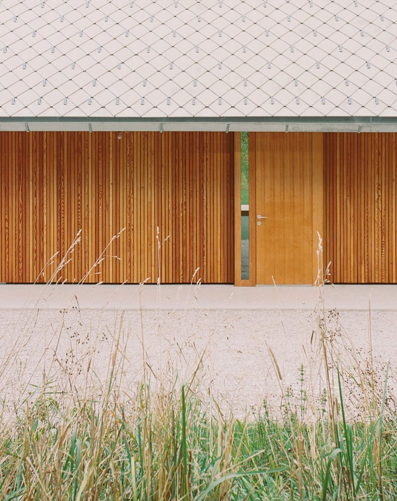 Timber-clad facade with flush door below grey shingled roof and concrete base edged by dry grasses