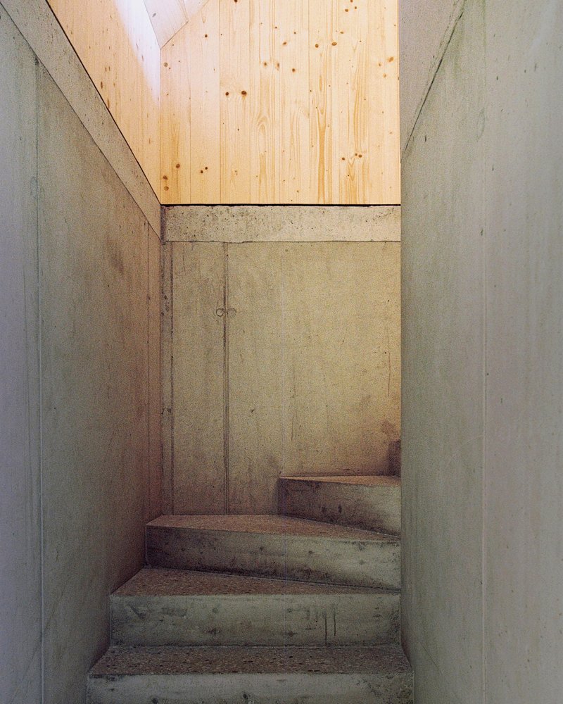 Narrow concrete stairwell with board-formed walls leading upward to a skylit plywood ceiling