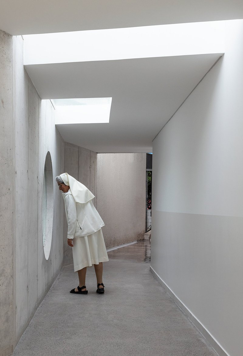 Skylit corridor with exposed concrete walls and a person standing beneath layered ceiling planes