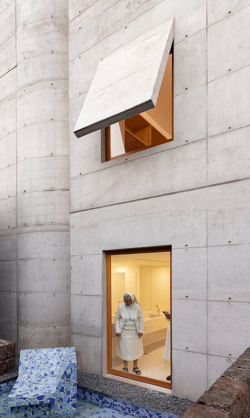 Board-formed concrete courtyard wall with two cantilevered window boxes and a person exiting at twilight