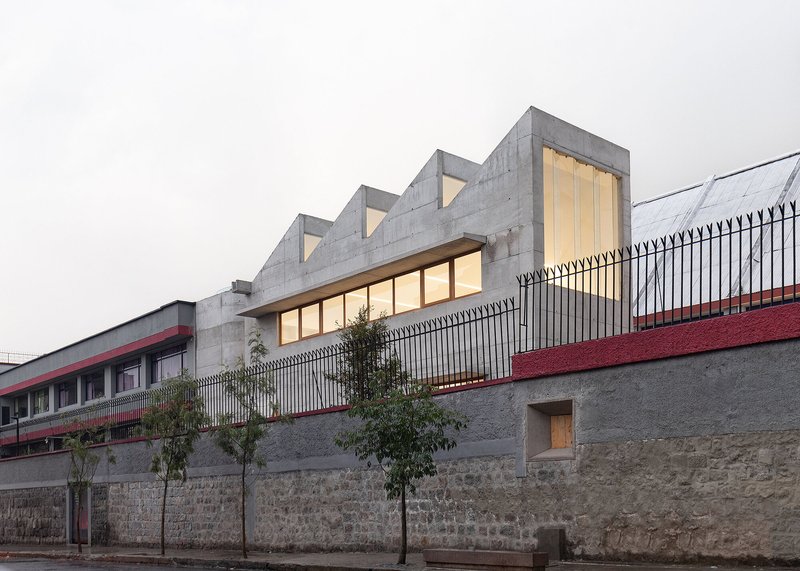 Street view showing the sawtooth roofline and illuminated interior behind black iron perimeter fence at twilight