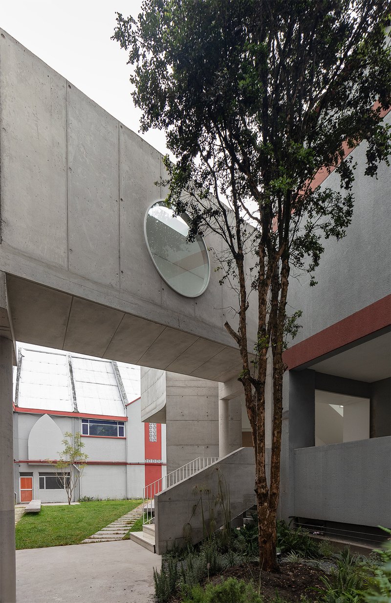 Courtyard view with concrete bridge overhead, circular mirror on the wall, and tree growing between volumes