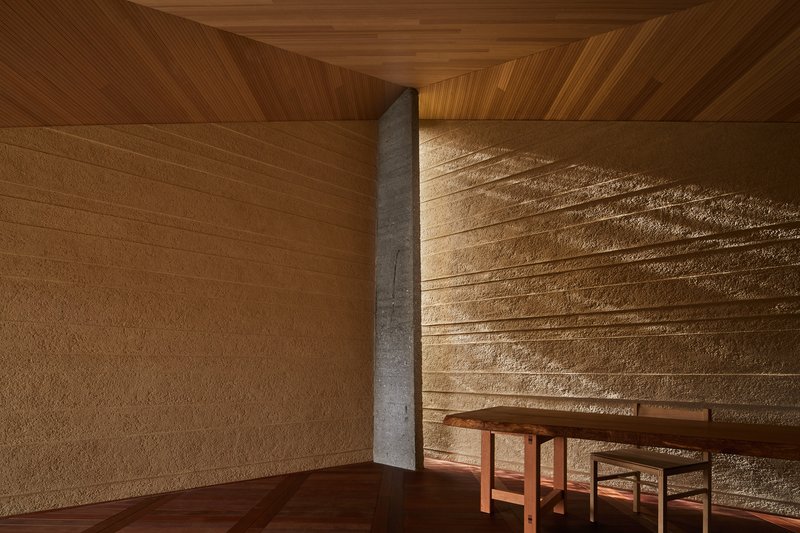 Timber seating below horizontally textured rammed earth wall with concrete column and wood ceiling