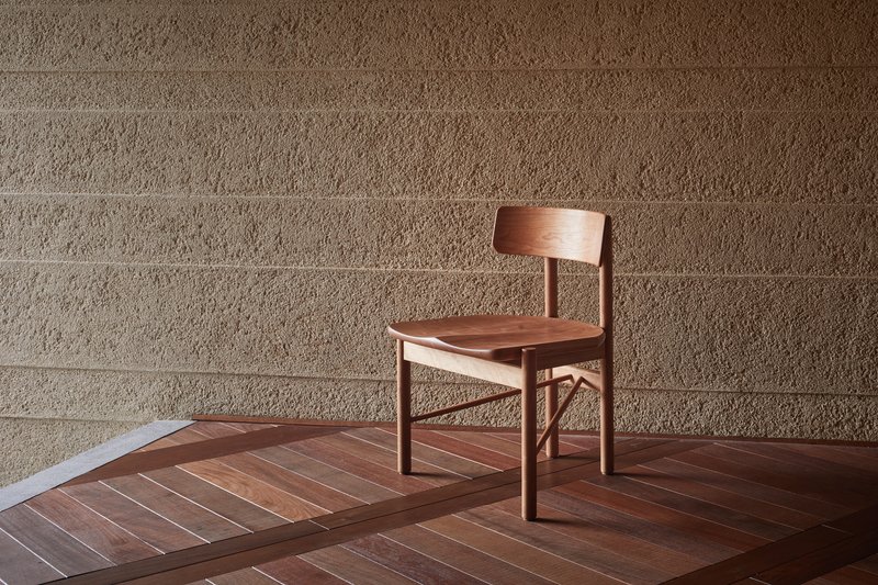 Timber chair on geometric wood floor against horizontal-grooved stone block wall in afternoon light