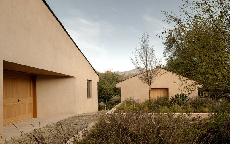 Gabled rammed earth volumes with timber doors opening onto a gravel courtyard with native plantings