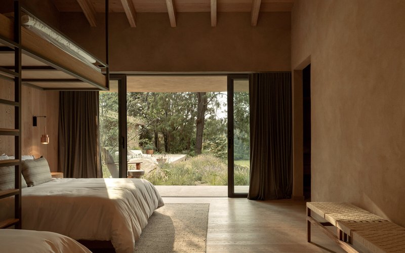 Bedroom with full-height glazing opening to a meadow under dappled morning sunlight