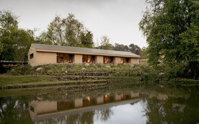 Long elevation of earth-toned building with regularly spaced window openings mirrored in pond water