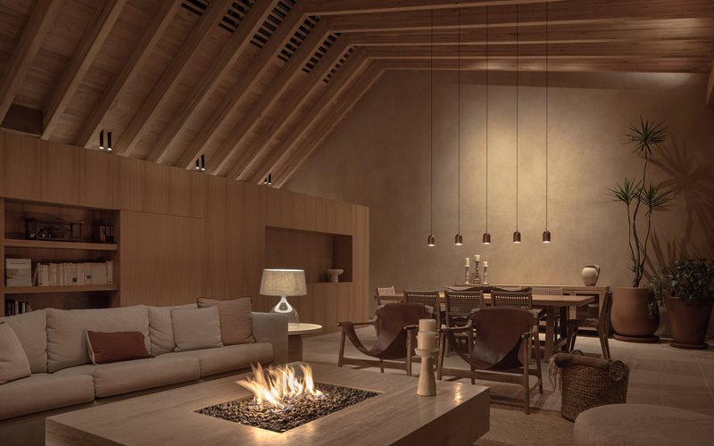 Open-plan living and dining space with exposed timber rafters and a central fire pit at dusk