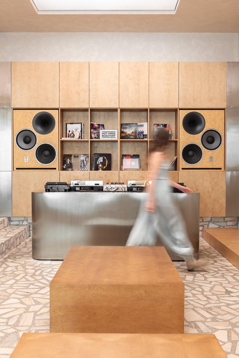 Plywood shelving wall with built-in circular speakers and a figure passing the brushed metal counter
