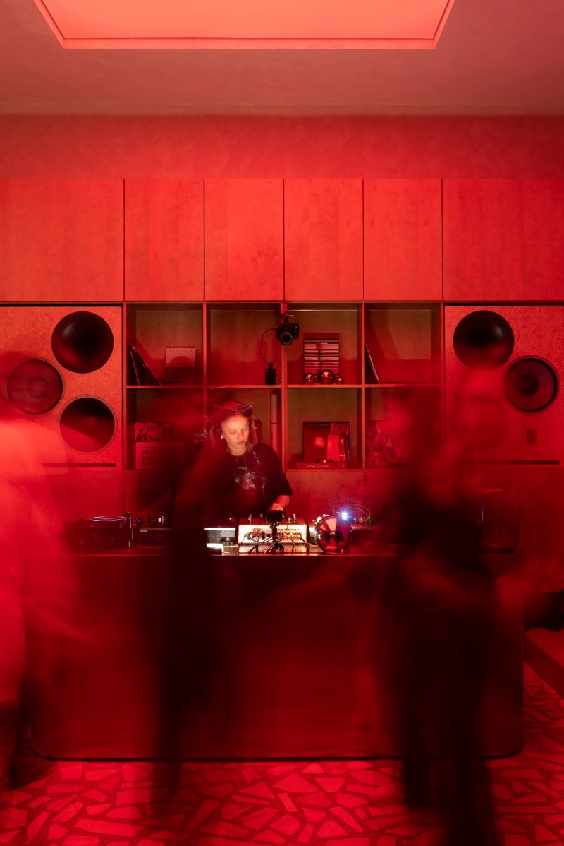 Red-lit service counter with circular openings and shelving units behind a figure working at dusk