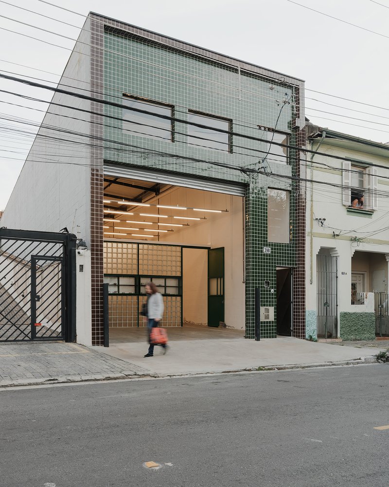 Street facade with green and brown tile cladding and large opening revealing illuminated interior as pedestrian passes