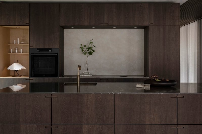 Kitchen island in dark timber with integrated sink facing a built-in oven and shelving niche