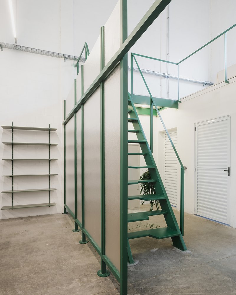 Double-height space with green steel staircase, glass railing above, and wall-mounted shelving under polished concrete floor