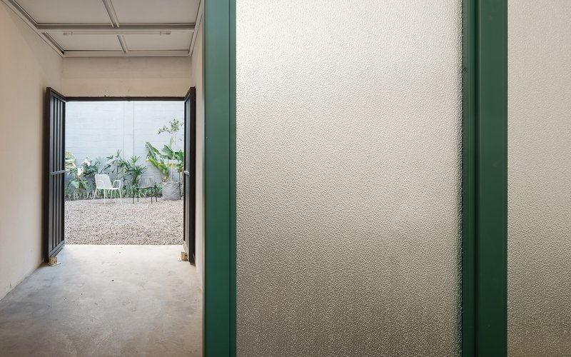 View through hallway with textured white wall trimmed in green steel toward courtyard with gravel and plants