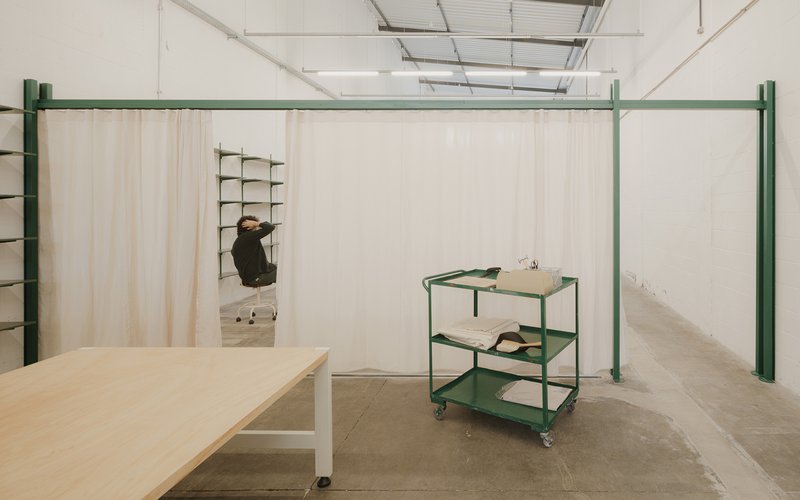 Workspace with green steel frame, fabric curtains, plywood table and rolling cart under exposed industrial ceiling