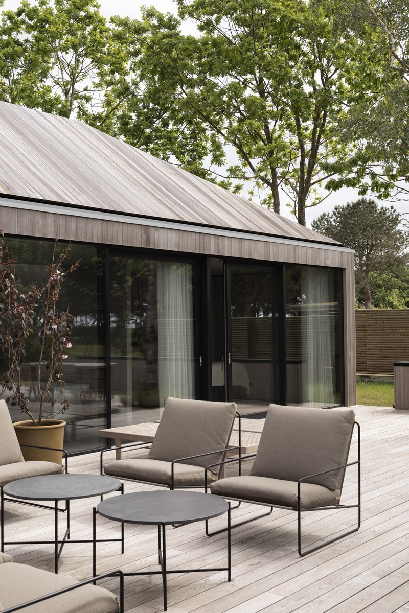 Outdoor seating area on pale wood decking with glass doors reflecting the surrounding green landscape