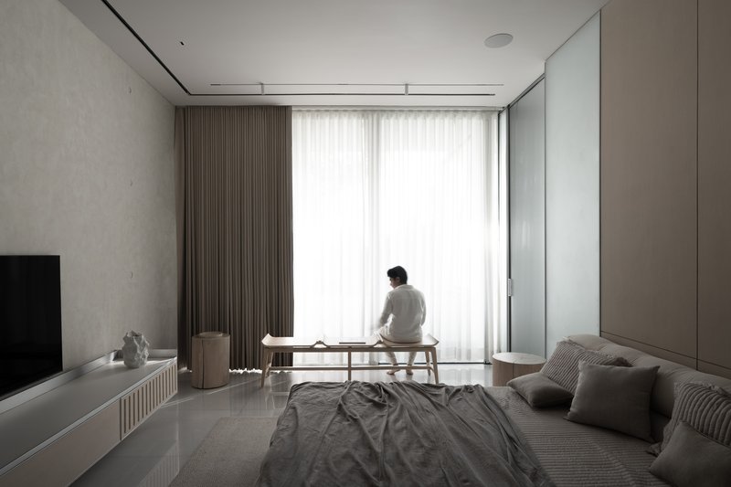Bedroom with white curtains drawn and a person seated at a low bench facing the window