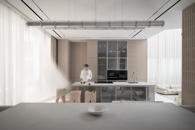 Open kitchen with exposed ducting overhead and a person standing at the island beneath sheer curtains