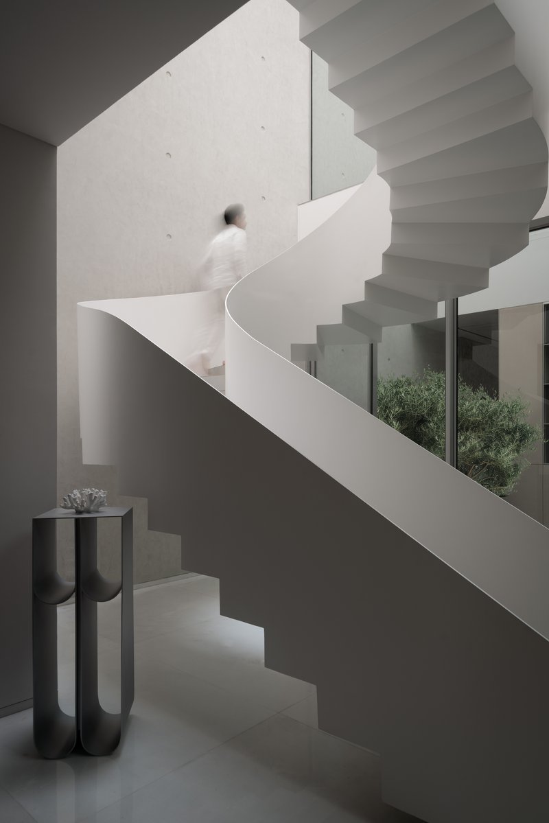 White sculptural staircase with a person ascending past a black pedestal and courtyard view