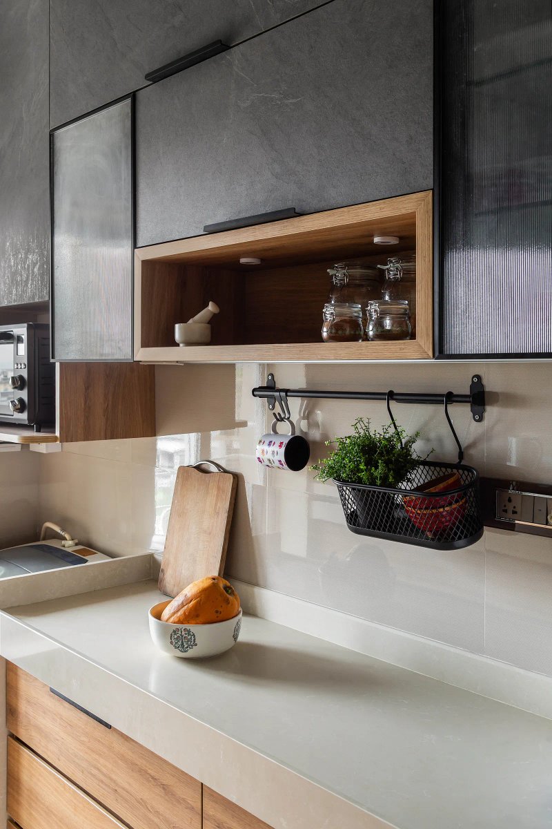 Concrete-like surface cladding, fluted glass and wood are the main ingredients of The Nest’s very contemporary kitchen. Two fluted shutters  wrapped in a black profile tuck in an open wooden niche completing the elevation. Anuja Kambli