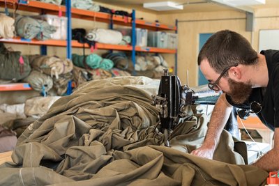 The image shows a man operating a vintage sewing machine, working on a large piece of khaki-colored 