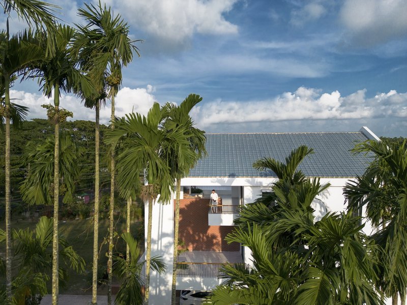 View of the white facade with orange brick accents and metal roof through tall palm trees