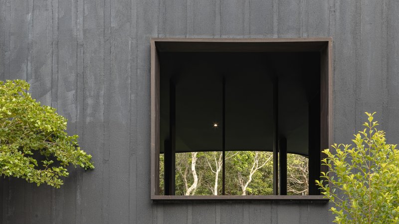 Deep-set square window punched into a charred-timber facade with leaves in the foreground