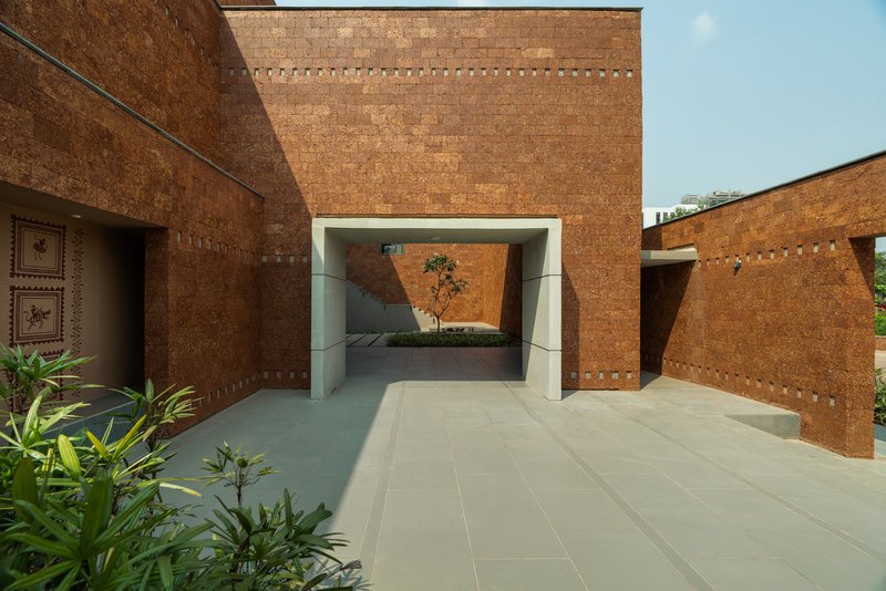 Red laterite volumes with a deep framed passage through a wall, leading to an inner courtyard