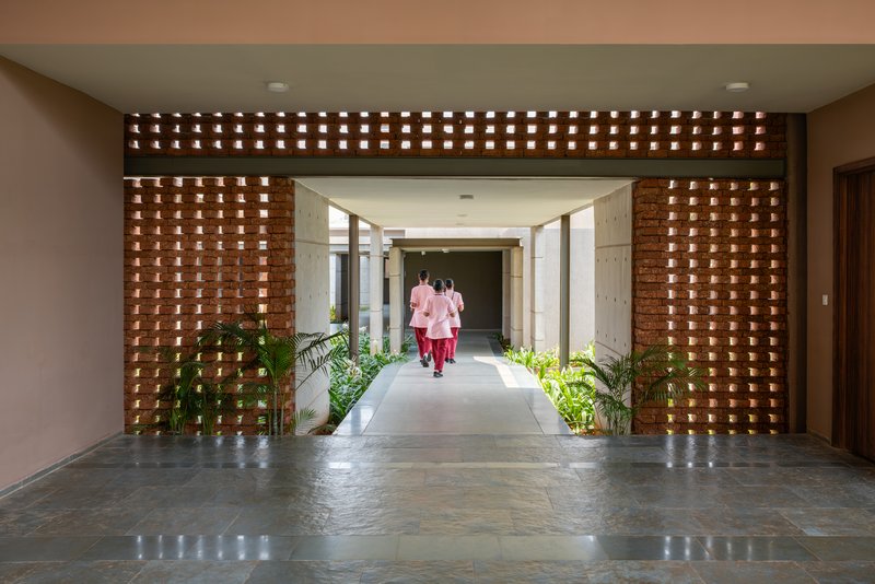 Entrance passage through a perforated laterite jali screen with nurses in pink walking toward a courtyard
