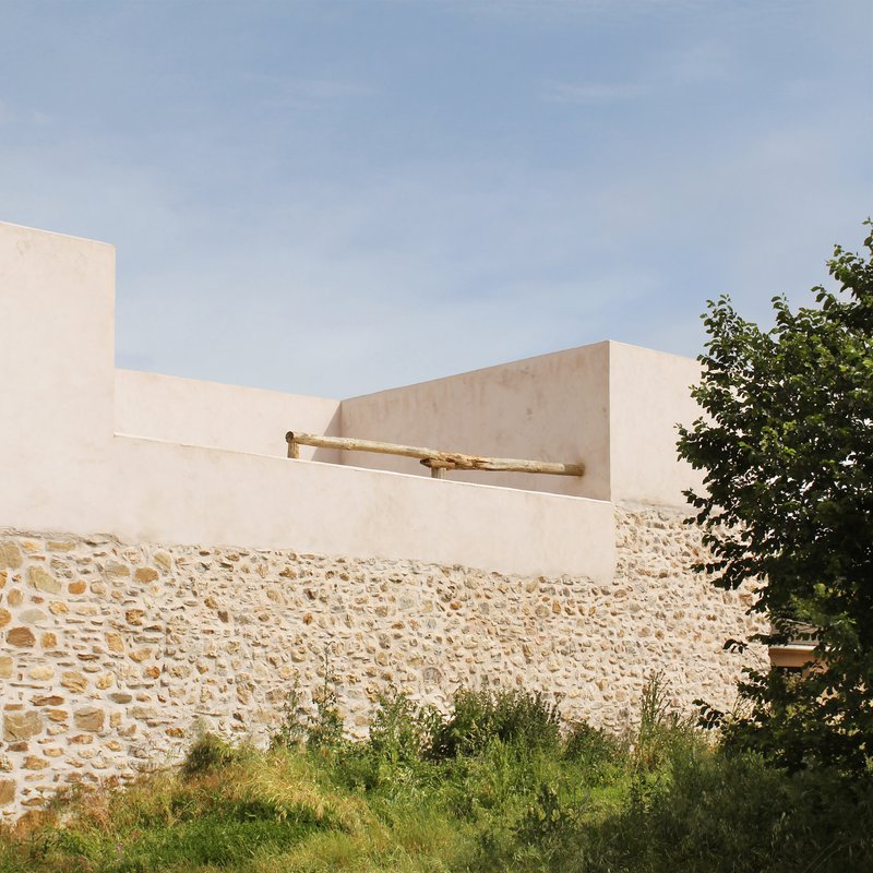 Detail of the white volume meeting the rough stone wall with a wooden roof beam projecting