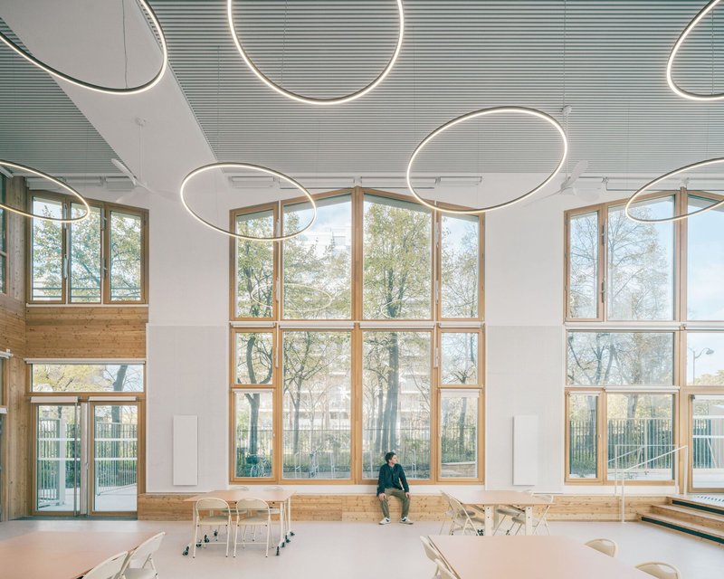Double-height community room with circular pendant light rings and a wall of timber-framed windows